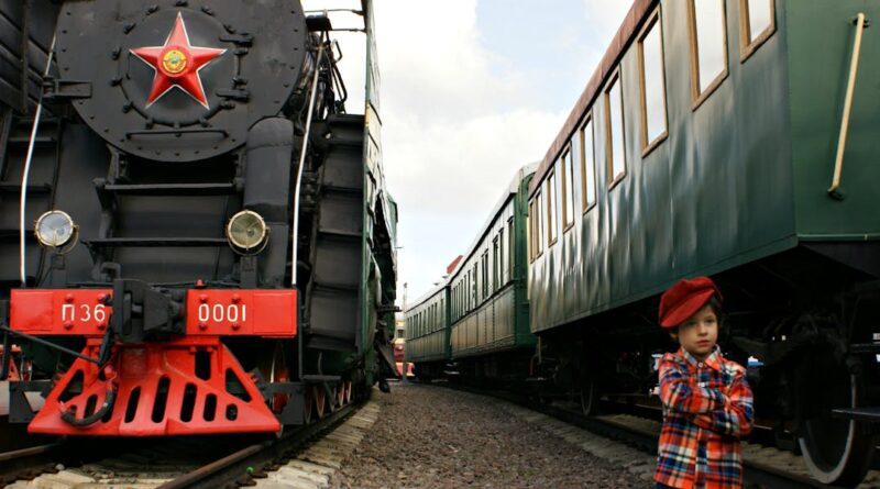 A child in a red hat stands by vintage trains on a railway track, showcasing travel history.
