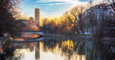 Scenic sunset view of Munich's riverside with a bridge, buildings, and trees reflecting in the water.