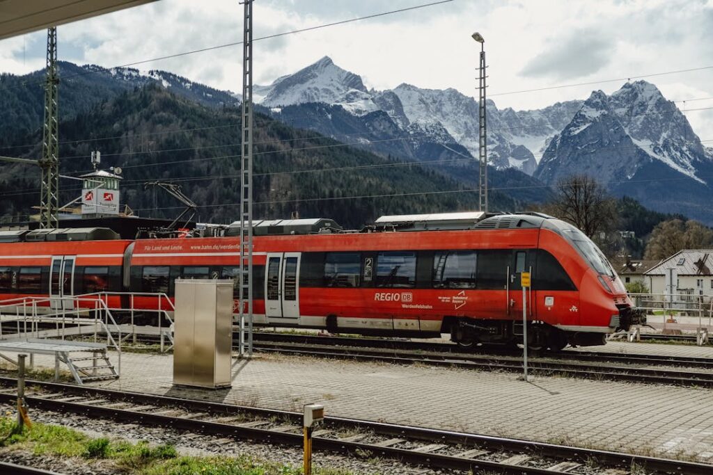 A red train at a scenic mountain station with snowy peaks in the background.