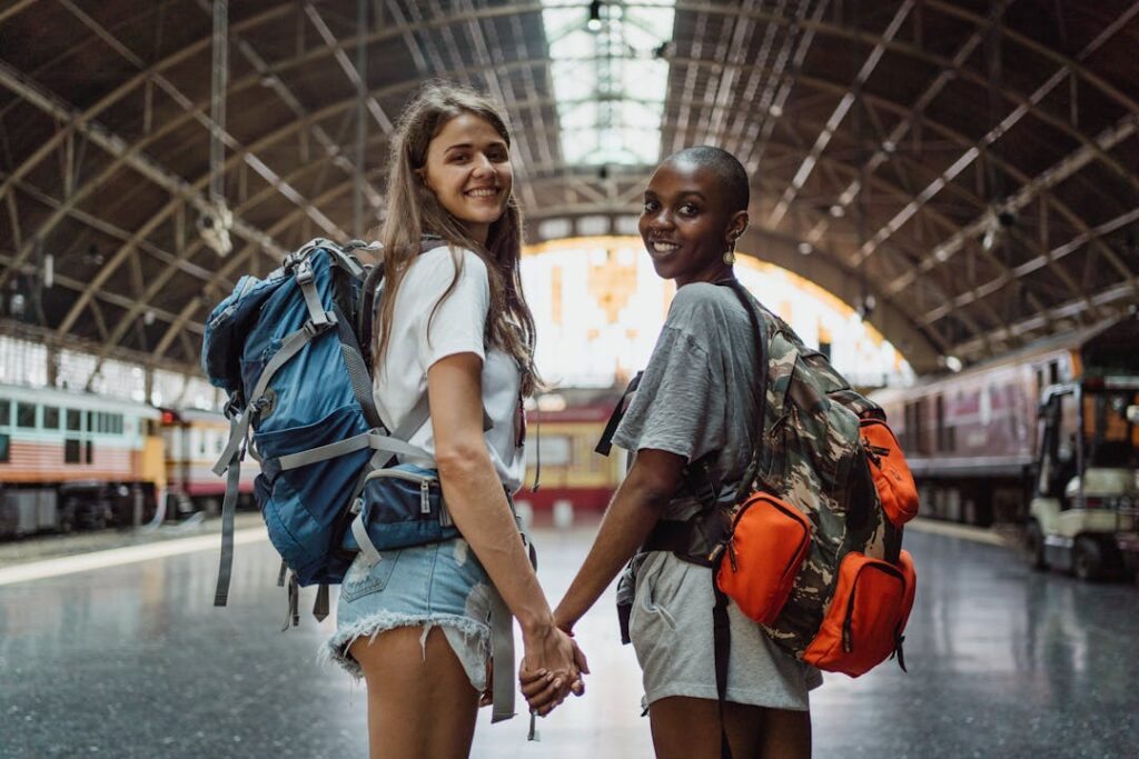 Two young women with backpacks holding hands, smiling at a railway station, ready for a travel adventure.