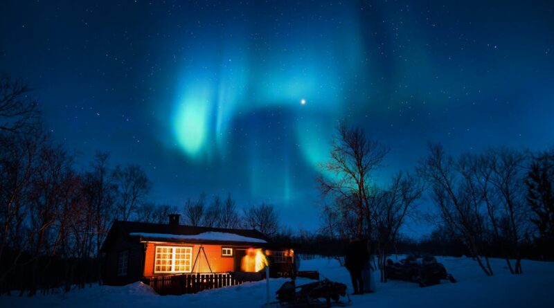 Cabin illuminated under a stunning aurora borealis and starry winter night sky.
